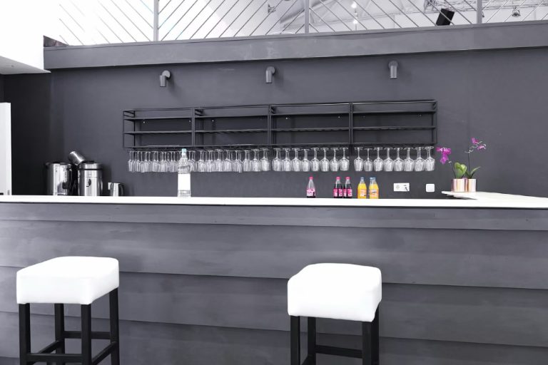 Echtes Foto: die lange Bar mit grauer Front, weißen Hockern und Gläserregal, ohne Gäste.