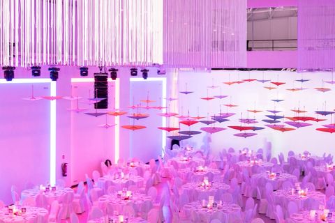 Echtes Foto: Gala-Dinner im Saal, runde Tische mit weißen Hussen, darüber bunte Schirme und Fadenleuchter in rosa Licht.