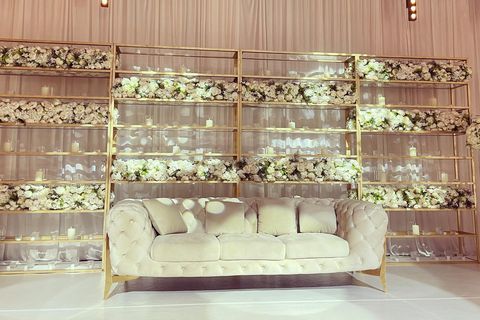 Echtes Foto: Weißes Chesterfield-Sofa vor einer goldenen Regalwand voller weißer Blumen.