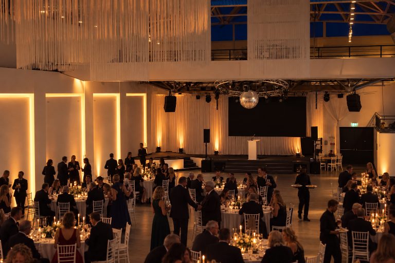 KI-Inszenierung, kein realer Anlass: Gala-Dinner mit Gästen in Abendgarderobe an runden Tischen bei Kerzenlicht, Rednerpult auf der Bühne.