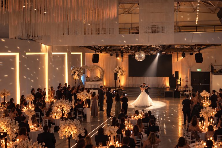 KI-Inszenierung, kein realer Anlass: Hochzeit am Abend, das Brautpaar tanzt im Lichtkegel den ersten Tanz, Gäste stehen am Rand der Tanzfläche, klatschen und filmen, andere sitzen an Tischen mit Kerzen.